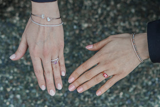 Two hands wearing stacked fine jewellery over a pebbled background, diamond tennis bracelets, a toi et moi bangle, bead bracelets, and pink gemstone rings.
