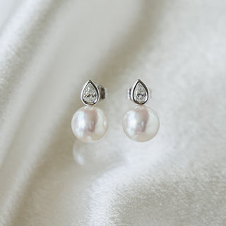 A pair of silver stud earrings featuring a small teardrop-shaped clear stone above a round white pearl, displayed on a soft white satin fabric background.