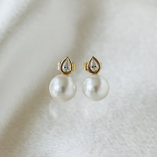 A pair of gold and pearl stud earrings with teardrop-shaped gold settings holding clear stones above large white pearl drops, displayed on a soft white textured surface.