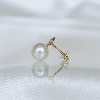 A single gold post earring with a round white pearl, shown on a soft white satin surface with a soft reflection.