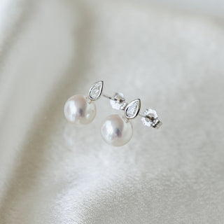 A pair of silver stud earrings featuring a marquise-cut clear stone and a round white pearl, displayed on a soft, textured white fabric background.