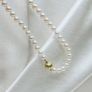 A white pearl necklace with a small gold ball charm resting on draped white satin fabric.