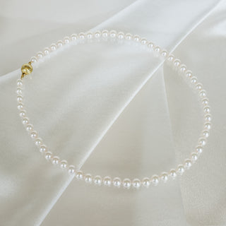 A single-strand pearl necklace with a gold clasp, arranged in a circle on draped white satin fabric.