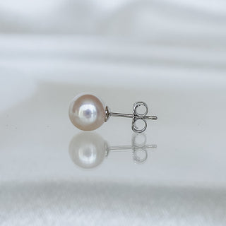 A single pearl stud earring with a silver post and butterfly back, resting on a reflective white surface against a soft white background.