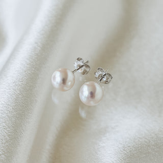 A pair of freshwater pearl stud earrings with silver butterfly backings, resting on a soft, draped white fabric background.