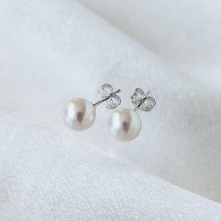 A pair of silver stud earrings with round white freshwater pearls, displayed on a soft, textured white fabric background.