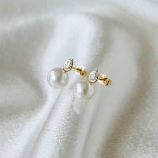 A pair of gold post earrings with a small marquise-cut clear stone and a dangling round white pearl, displayed on a soft white satin fabric background.