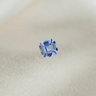 A single loose emerald-cut light blue sapphire, centered against a soft, warm beige textured background.