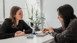 Two women sitting at a table with jewelry, discussing items.