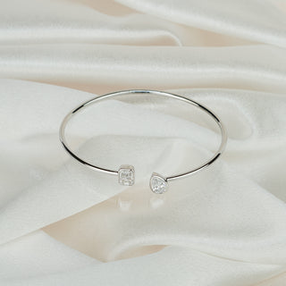 Open white gold bangle with bezel set lab grown diamonds lying on ivory satin, emerald cut and pear shape at the gap.