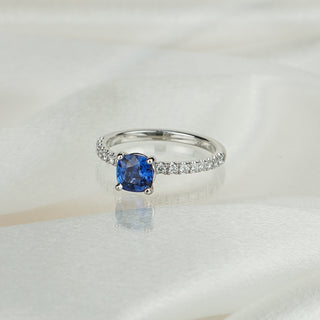 Cushion cut blue Sapphire engagement ring in white gold on satin fabric, four-prong center with pavé Diamonds along the shank.