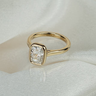 Cushion cut lab grown diamond bezel engagement ring in yellow gold on satin fabric, full bezel above a cathedral shank.