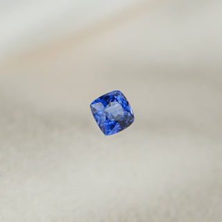 A single loose cushion-cut blue gemstone, likely a sapphire, centered against a soft, out-of-focus light beige background.
