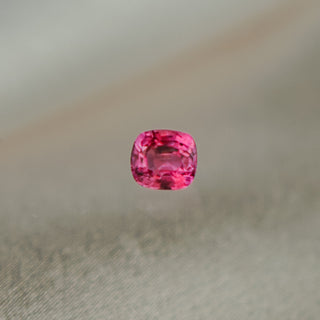 A cushion-cut pink gemstone shown in a loose stone format against a soft-focus gray textured background.