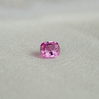 A single cushion-cut pink gemstone, likely a sapphire, resting on a textured light gray fabric surface.