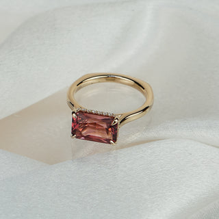 Pink Tourmaline hidden halo engagement ring in yellow gold on satin fabric, radiant cut center set east-west over concealed Diamonds.