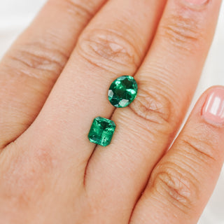 Two green gemstones, one oval and one rectangular, displayed on a person's finger against a light background.