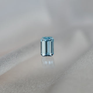 A single rectangular emerald-cut light blue aquamarine, centered on a soft, draped light gray fabric background with a subtle reflection below.