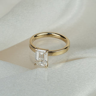Emerald cut lab grown diamond solitaire in yellow gold on satin fabric, four-prong center on a rounded pipe shank.
