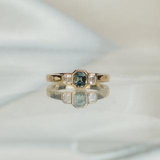 Yellow gold three-stone engagement ring with an emerald cut teal Sapphire center and Asscher cut Diamonds in bezels.