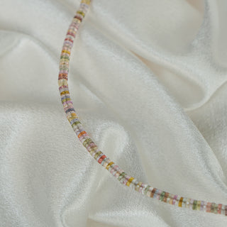 Multi-color Sapphire tire bead strand draped on ivory satin, flat-sided beads catching light along the curve.