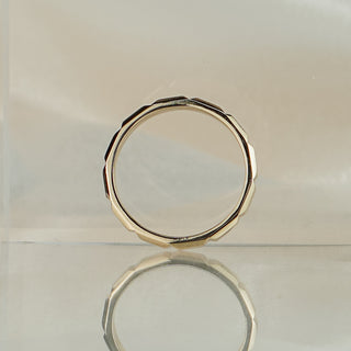 Faceted yellow gold wedding band standing upright with reflection, showing the paneled exterior and round interior.
