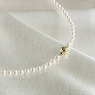 A delicate pearl necklace with a polished gold bead accent, resting on draped white satin fabric.