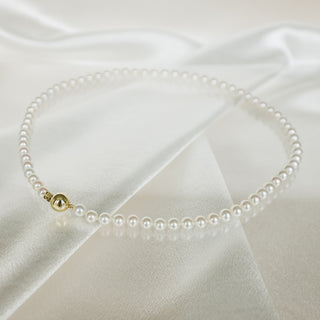 A delicate pearl necklace with a small gold bead accent, displayed on draped white satin fabric.