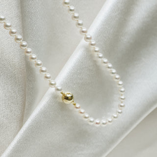 A close-up of a pearl necklace draped on white satin fabric, featuring a single polished gold bead accent.