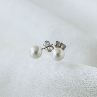 A pair of stud earrings featuring round white freshwater pearls set in silver-tone metal, resting on a soft, textured white fabric surface.