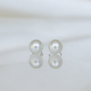 A pair of round white freshwater-style pearl stud earrings with silver posts, displayed on a reflective white surface against a soft white background.