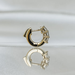 A single gold huggie hoop earring with a basket-like cluster of clear stones on the front, displayed on a reflective white surface against a soft white background.