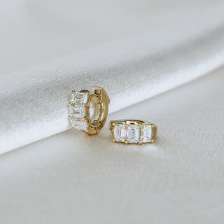 A pair of gold huggie hoop earrings with baguette-cut clear crystals, displayed on a soft white satin fabric background.