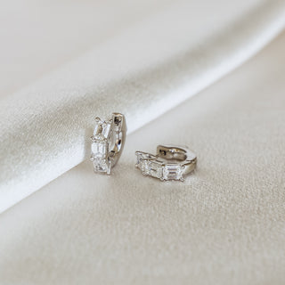 A pair of silver huggie hoop earrings with baguette-cut clear crystals, displayed on a soft, draped white satin fabric.