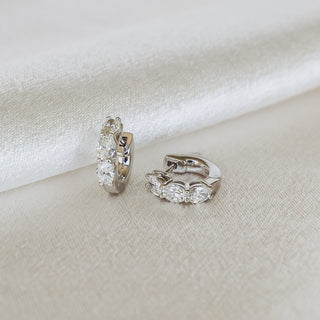 A pair of small silver huggie hoop earrings with clear marquise-cut gemstones, displayed on a textured cream fabric background.