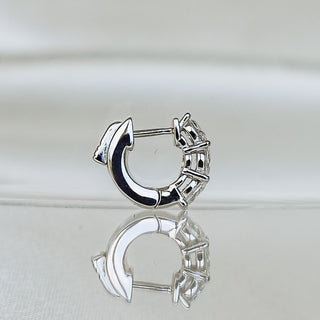 A polished silver huggie hoop earring with a small decorative clasp, shown on a reflective light gray surface against a soft light gray background.