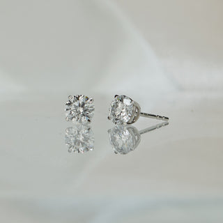Lab grown diamond round brilliant stud earrings in white gold, four-prong settings with reflection on a pale surface.