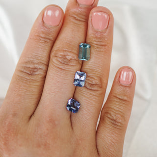 Three rectangular gemstones in different shades of blue and gray are arranged vertically on a person's finger against a soft white fabric background.