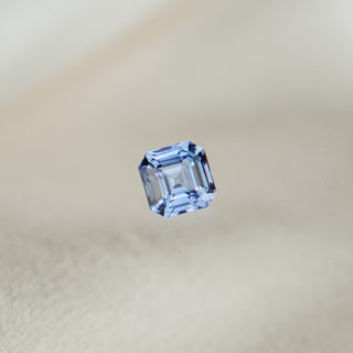 A single square-cut light blue sapphire, centered against a soft, out-of-focus beige background.