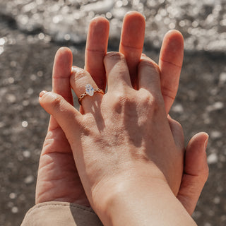 Close-up of two hands with a marquise engagement ring, blurred water background