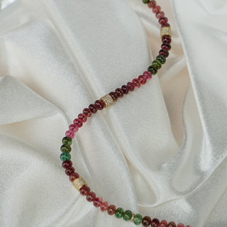 Multi-color Tourmaline bead strand draped on ivory satin, oval beads shifting from pink to green along the curve.