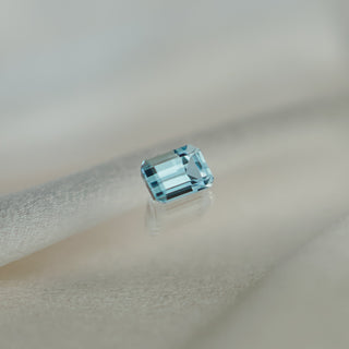 A single rectangular emerald-cut light blue aquamarine, displayed on a soft beige fabric surface with a subtle reflection.