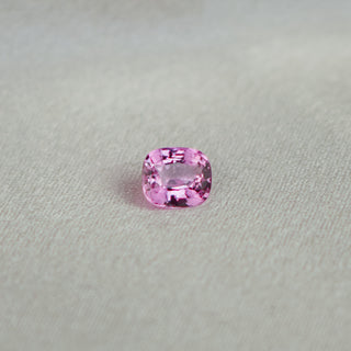 A single cushion-cut pink gemstone, likely a sapphire, resting on a textured light gray fabric surface.