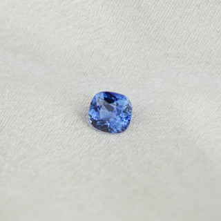 A cushion-cut blue sapphire gemstone displayed on a textured light gray fabric background.