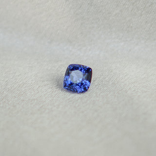 A cushion-cut blue sapphire gemstone with brilliant facets, displayed on a textured light gray background.
