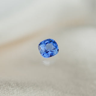 A single cushion-cut blue gemstone, likely a sapphire, shown on a soft, light beige fabric surface with a subtle reflection.