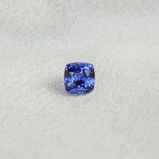 A cushion-cut blue sapphire gemstone with brilliant sparkle, centered on a textured light gray fabric background.