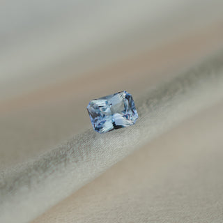 A single rectangular emerald-cut light blue sapphire, resting on a soft beige fabric surface.