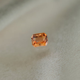 A single rectangular emerald-cut orange gemstone, likely a sapphire or moissanite, centered on a soft, textured light beige fabric background.