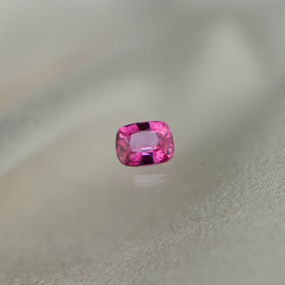 A small rectangular cushion-cut pink gemstone with faceted edges, photographed on a soft light gray surface with a faint reflection below.